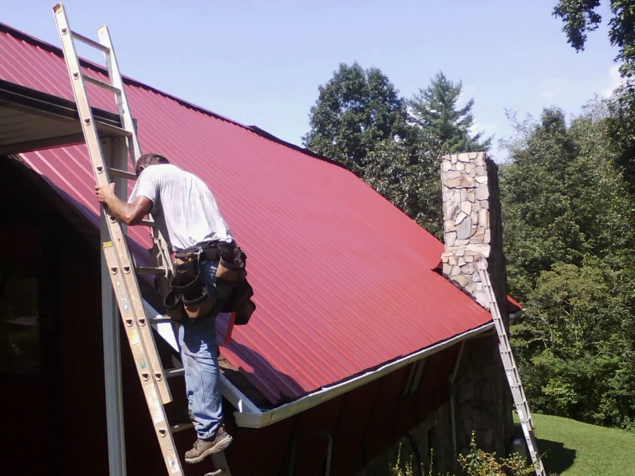 Roofer on a ladder beside a red metal roof and stone chimney — Soffit & Fascia Repair by Roofing Brothers in Bellmore