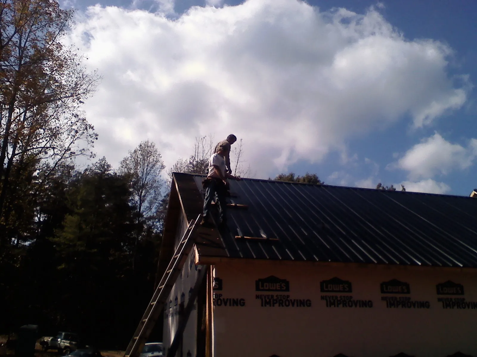 Roofer fastening dark metal panels high against a cloudy sky, from a Roof Inspection job in Bellmore